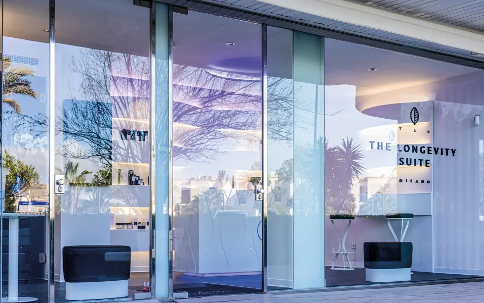 Vista esterna di un centro The Longevity Suite con ampie vetrate e logo visibile, riflessi degli alberi nelle finestre